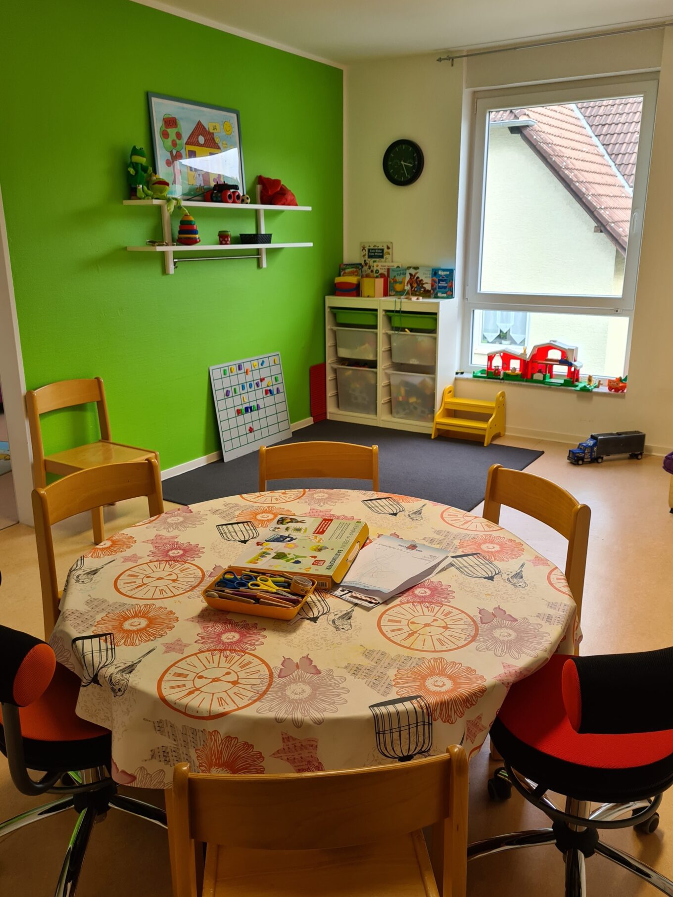 Ein Spielzimmer mit einem runden Tisch mit Bastelzubehör, einem Spielzeugregal und einem Spielteppich